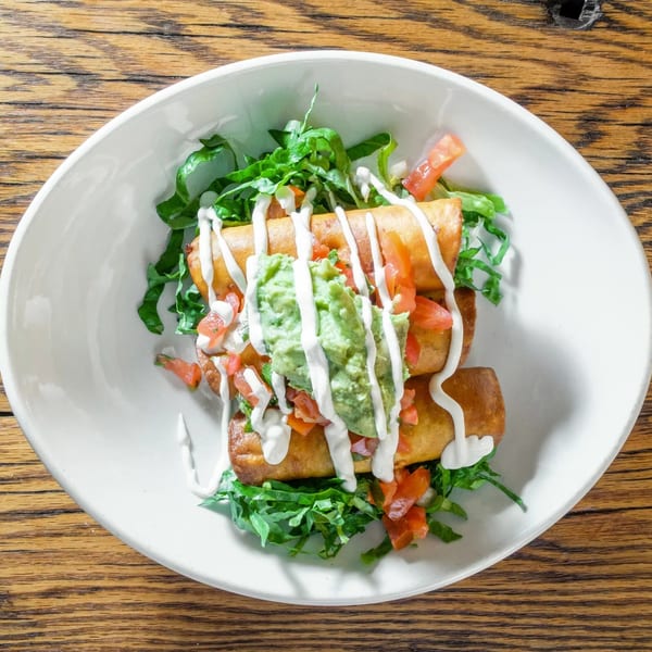 Golden taquitos topped with guacamole, sour cream, diced tomatoes, and lettuce in a white bowl.