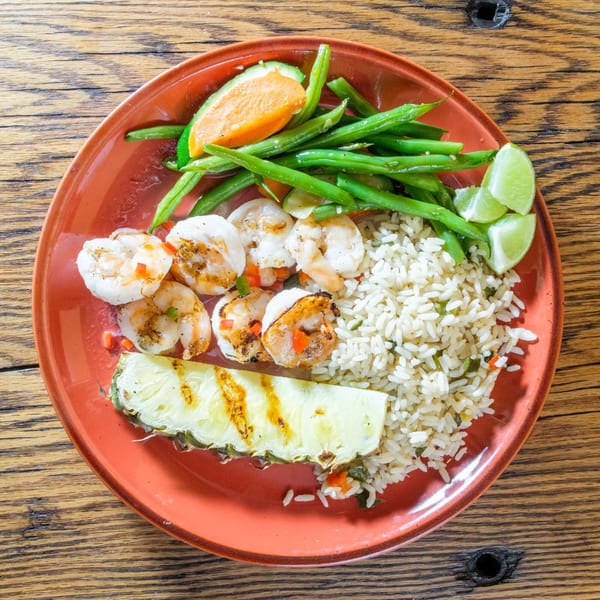 Grilled shrimp, rice, green beans, grilled pineapple, and lime wedges on a red plate.