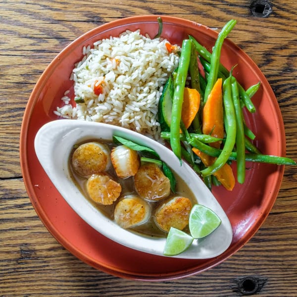 Seared scallops in sauce with lime wedges, rice, green beans, and carrots on a red plate.