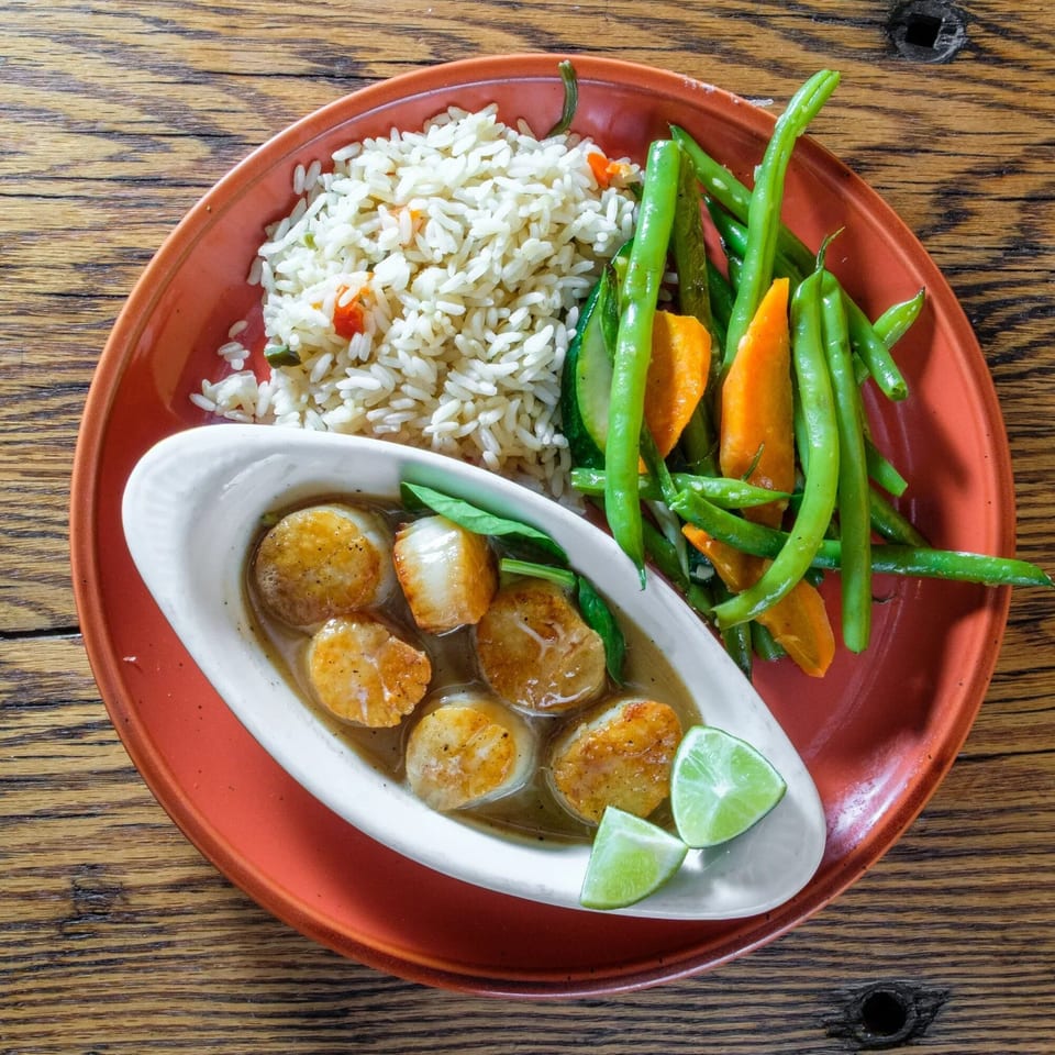 Seared scallops in sauce with lime wedges, rice, green beans, and carrots on a red plate.